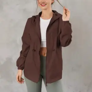 Frau in brauner Jacke mit Kapuze und Kordelzug an der Taille vor Betonwand