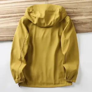 Senfgelbe Softshelljacke mit Kapuze von hinten, am Saum mit Zugband, vor Holzwand