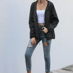 Frau in kurzer schwarzer Regenjacke mit Kapuze, weißem Top und Jeans an einer hellen Wand