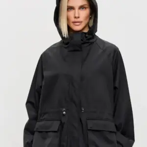 Frau in schwarzer kurzer Jacke mit Kapuze, Taillenzug und großen Taschen vor hellem Hintergrund